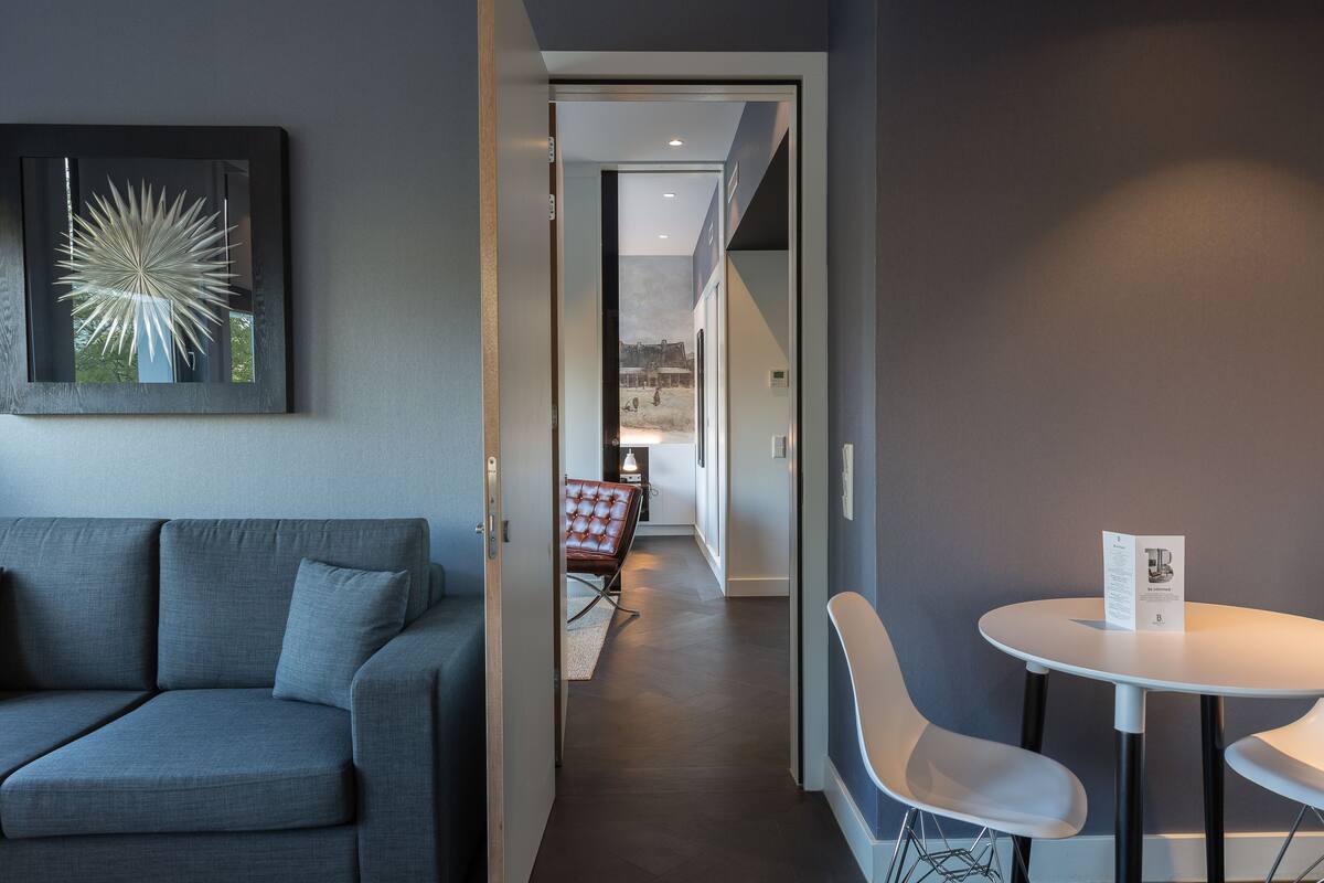 A modern hotel room with a grey sofa, a small dining table with two chairs, and a framed artwork on the wall.