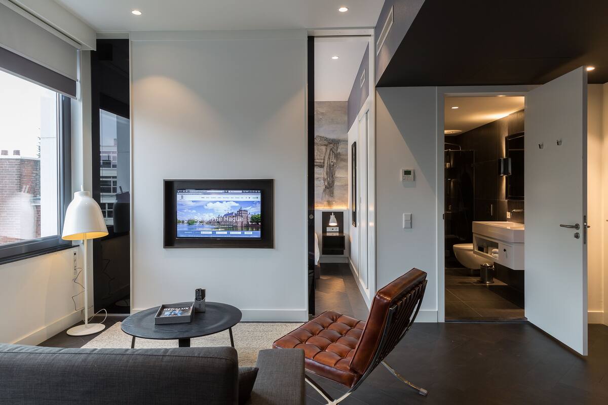 A modern hotel room with a large window, a flat-screen TV mounted on the wall, a round table with a lamp, a leather armchair, and a view of the cityscape.