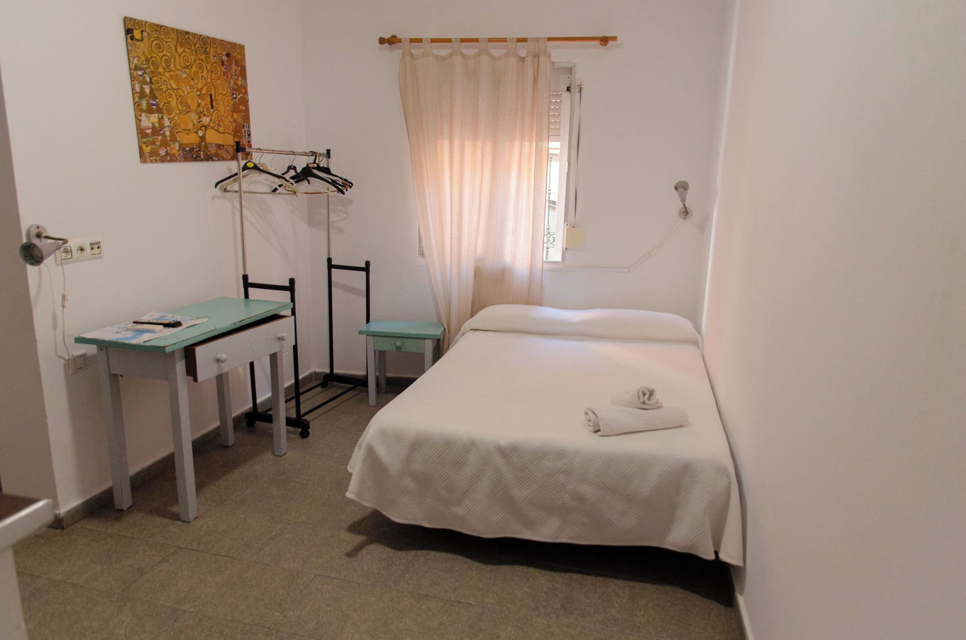 A small, clean room with a bed, a desk, and a chair. There is a curtain on the window and a painting on the wall.