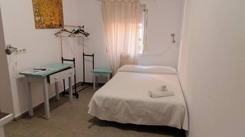 A small, clean room with a bed, a desk, and a chair. There is a curtain on the window and a painting on the wall.