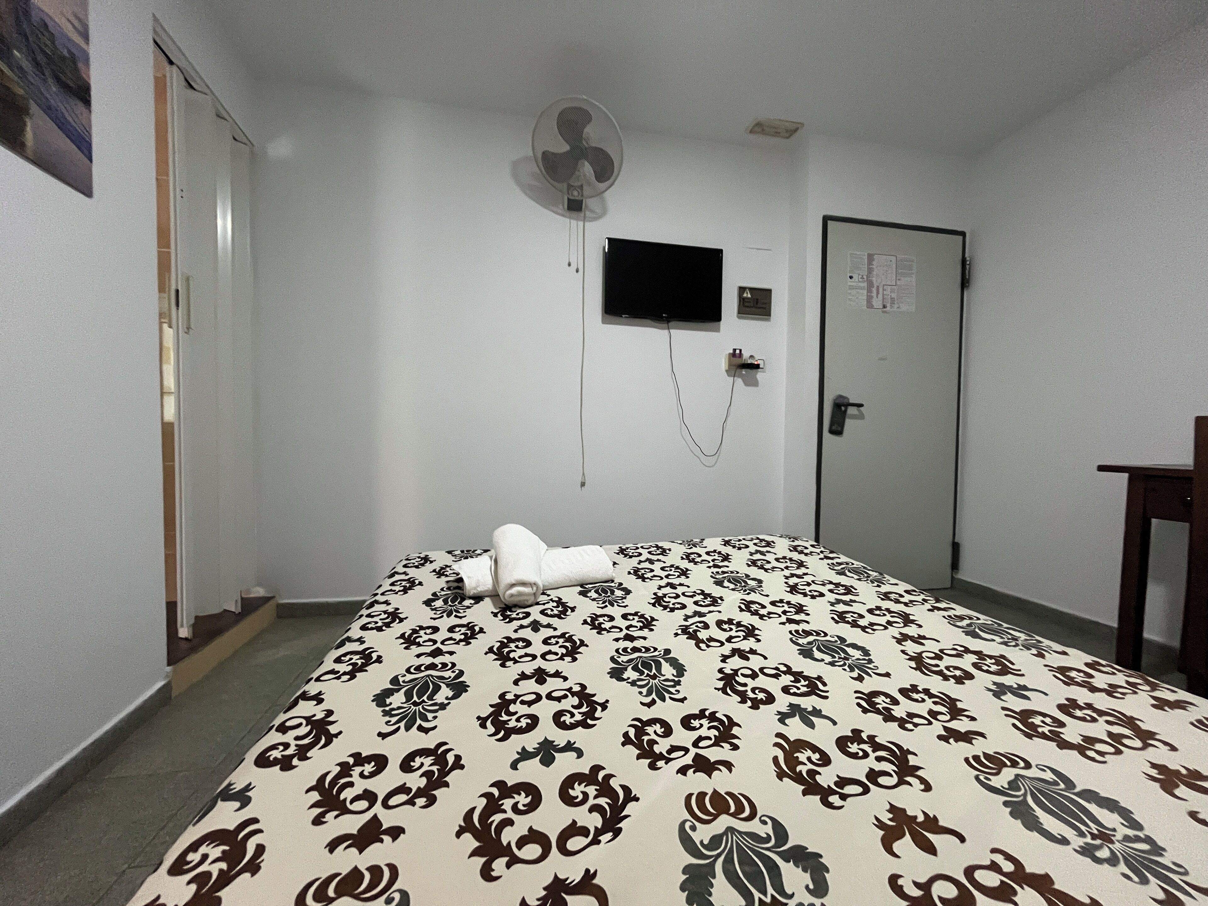 Una habitación de hotel con cama, televisión instalada en la pared, una puerta y un ventilador de pared.