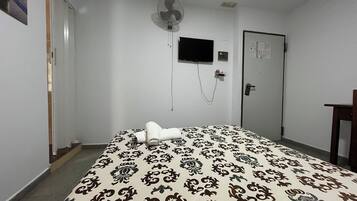 Una habitación de hotel con cama, televisión instalada en la pared, una puerta y un ventilador de pared.