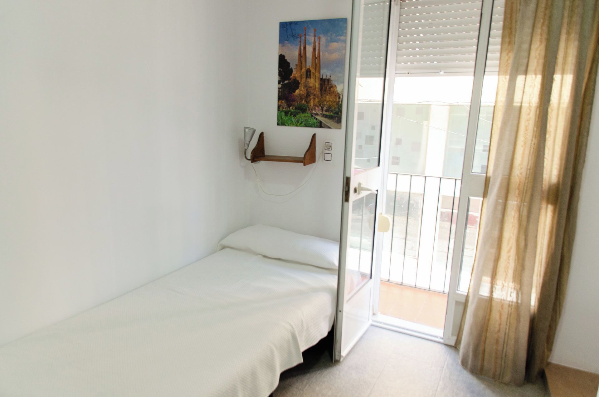 Una habitación pequeña con cama individual, una cama blanca, un teléfono en la pared y una imagen de una catedral.