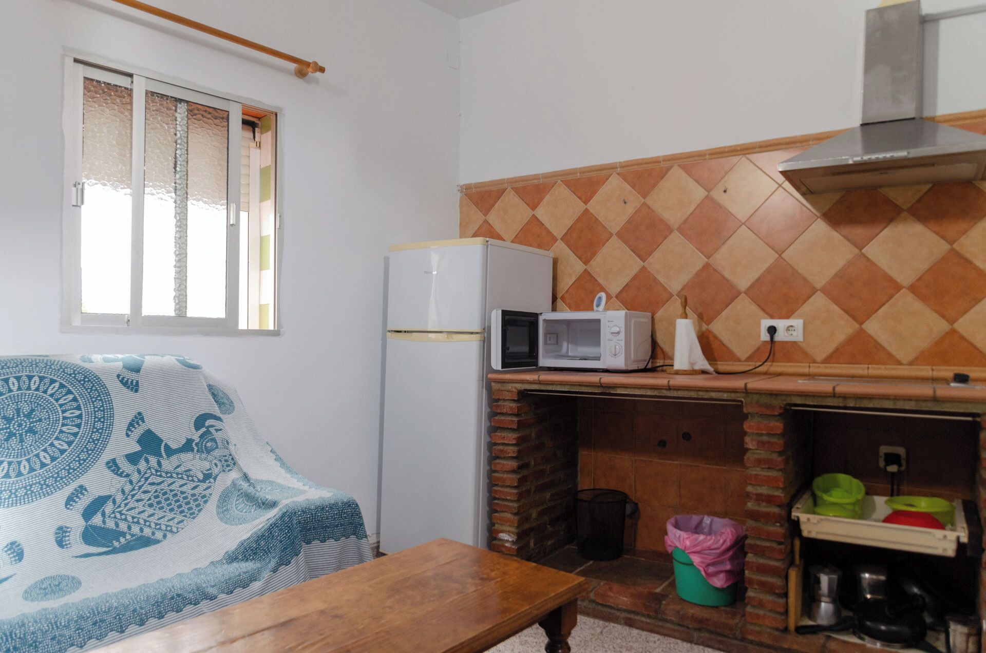 Una cocina compacta con refrigerador, microondas y horno de ladrillo. Hay una ventana con persianas, un sofá estampado y una mesa de madera.