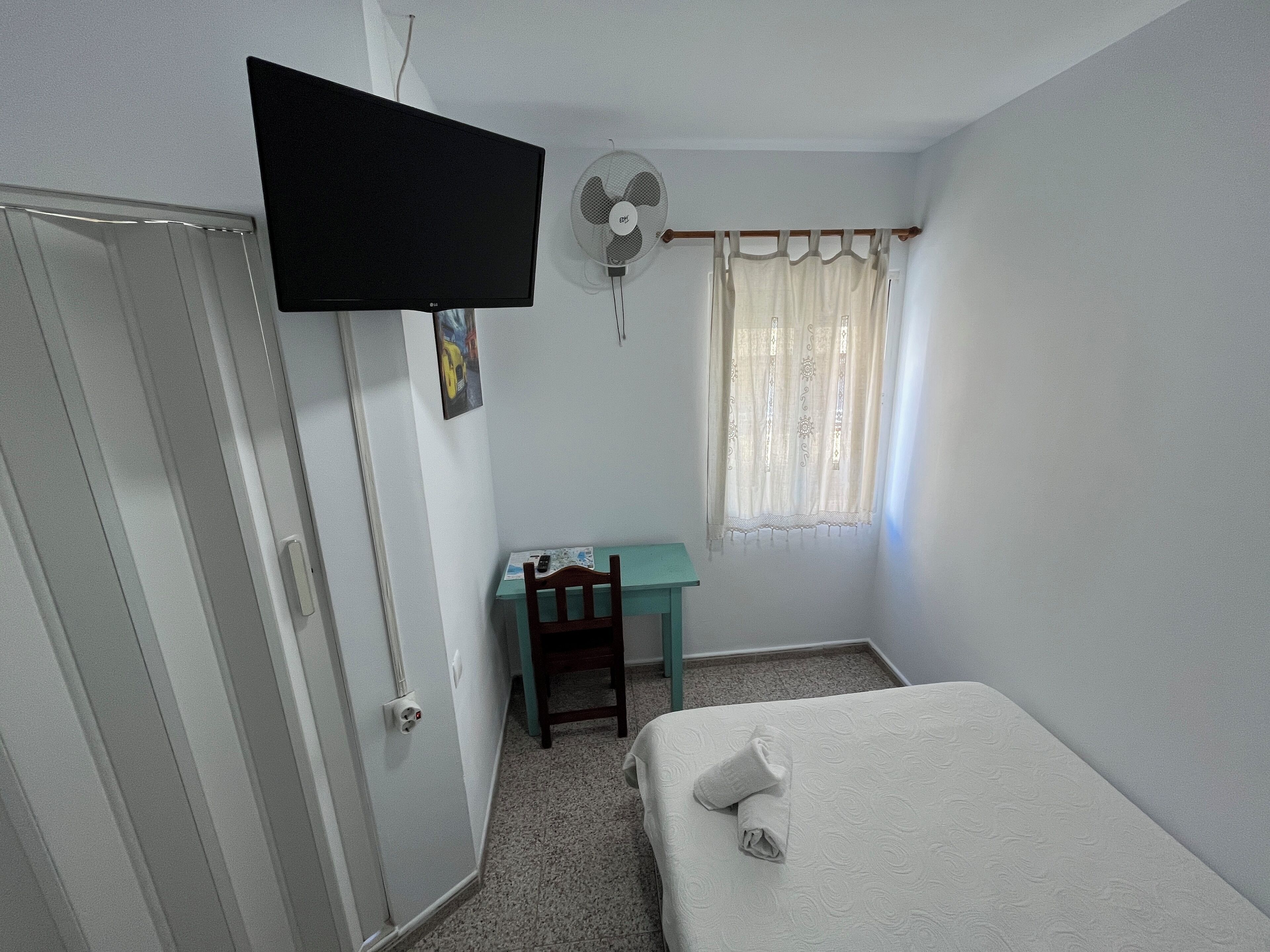 A bedroom with a bed, a desk, a chair, a TV, and a window with curtains.