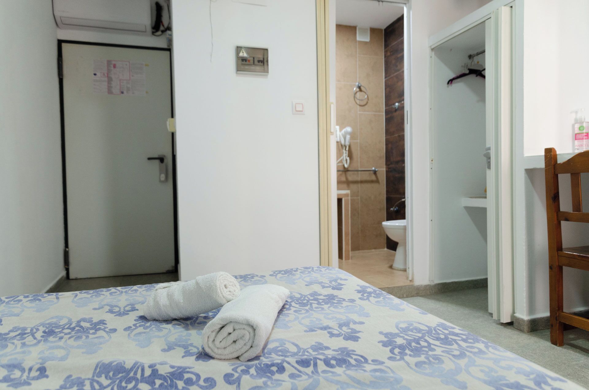 A hotel room with a bed, a wooden chair, a bathroom with a shower, and a toilet visible through an open door.