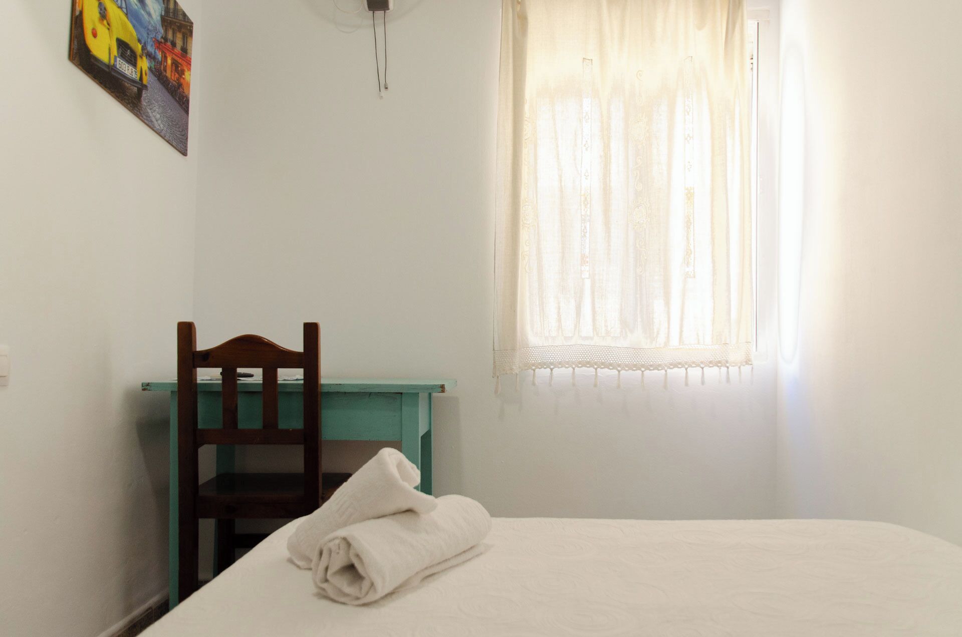 Una habitación con una cama, una silla de madera, una mesita pequeña, un cuadro en la pared y una ventana con cortinas traslúcidas.