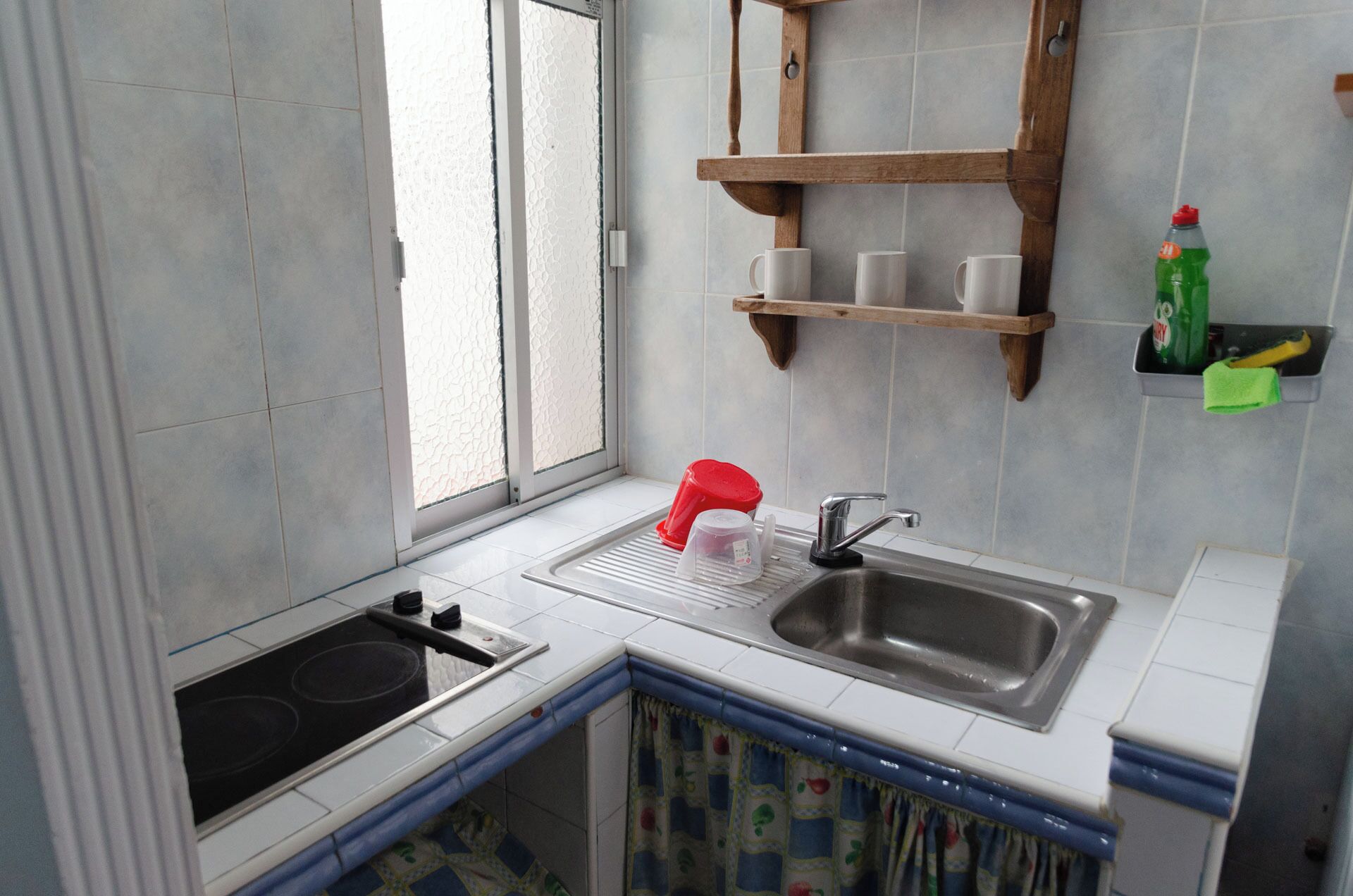 Una cocina con un lavamanos, estufa y ventana. Hay estantes sobre el lavamanos y una botella a la derecha.