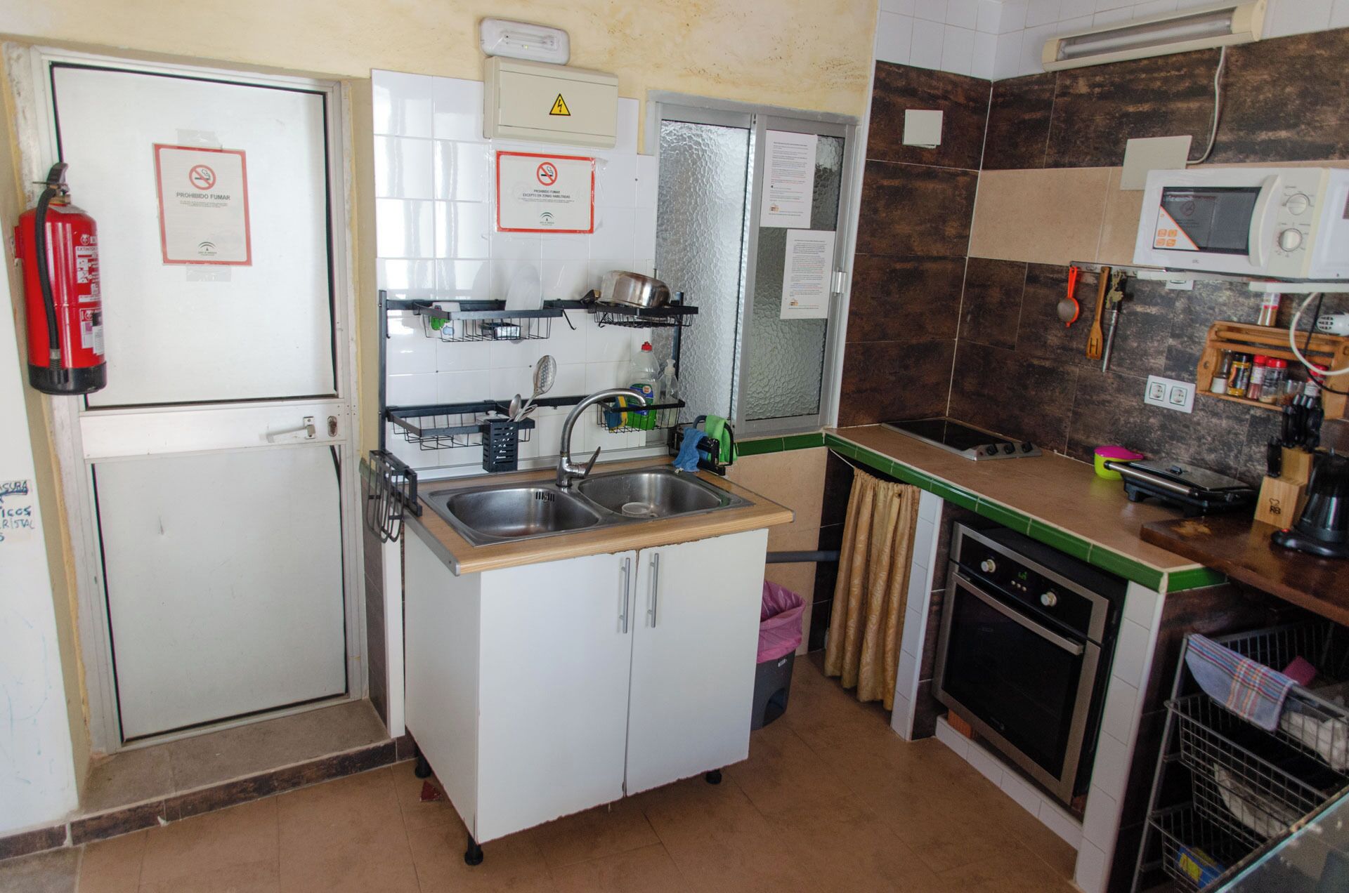 A compact kitchen with a sink, stove, and microwave. There are signs indicating no smoking and a fire extinguisher.