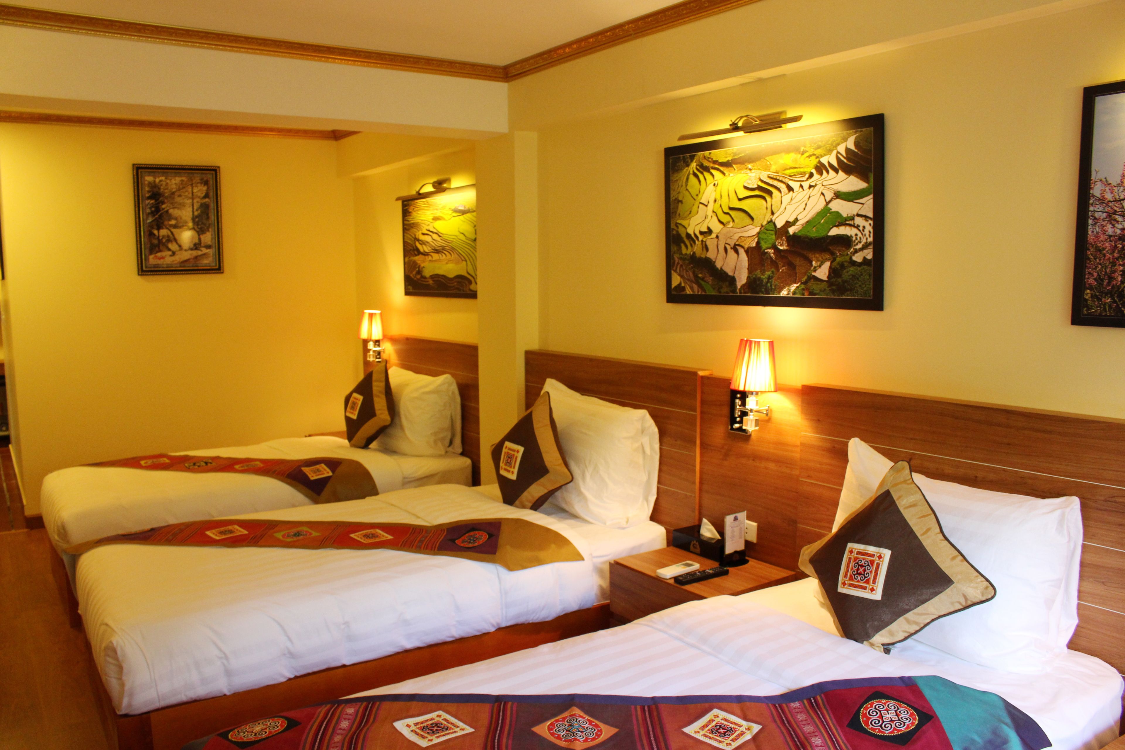 A hotel room with two beds, each with white linens and decorative pillows. There are wall-mounted lamps and framed artwork above the beds.