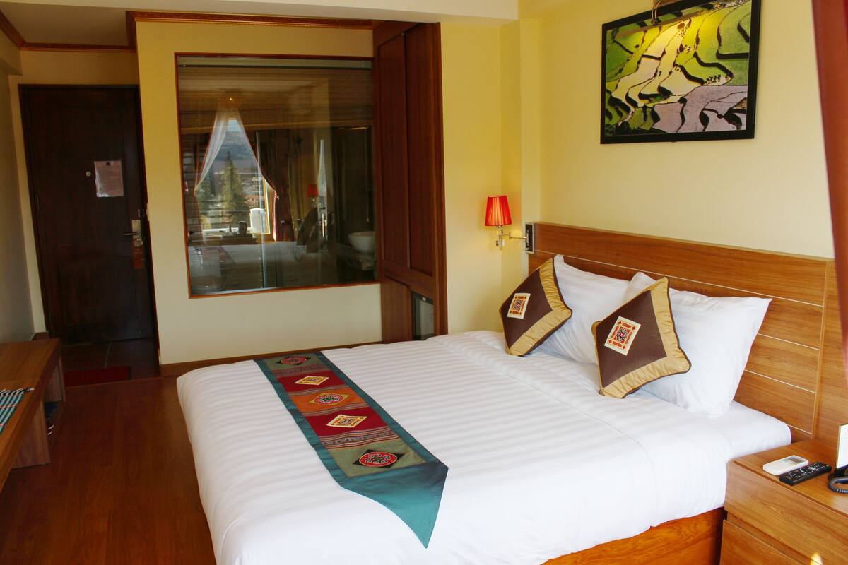 Habitación de hotel con una cama bien tendida, cabecera de madera, mesita de noche con lámpara y un cuadro en la pared.