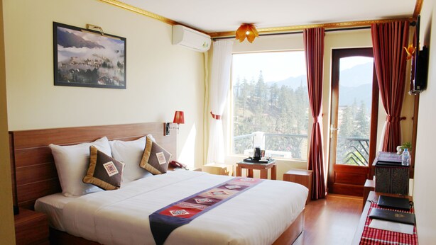 Quarto de hotel com uma cama grande, uma mesinha, uma cadeira, uma janela com cortinas e um quadro na parede.