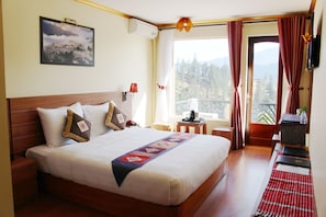 Habitación de hotel con una cama grande, una mesita pequeña, una silla, una ventana con cortinas y un cuadro en la pared.