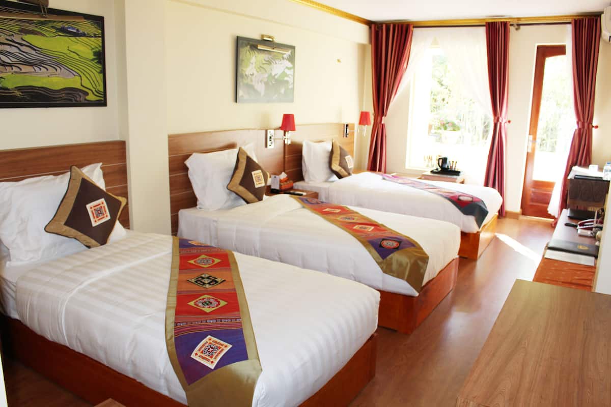 Habitación de hotel con tres camas, cada una con un edredón de diferente estampado. Hay dos lámparas de pared, un cuadro en la pared y una ventana con cortinas.