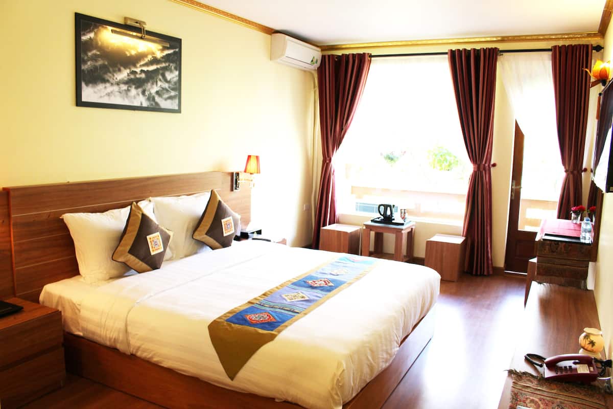 Habitación de hotel con una cama grande, una mesita de noche, una lámpara, aire acondicionado, una ventana con cortinas y una pequeña mesa con un hervidor.