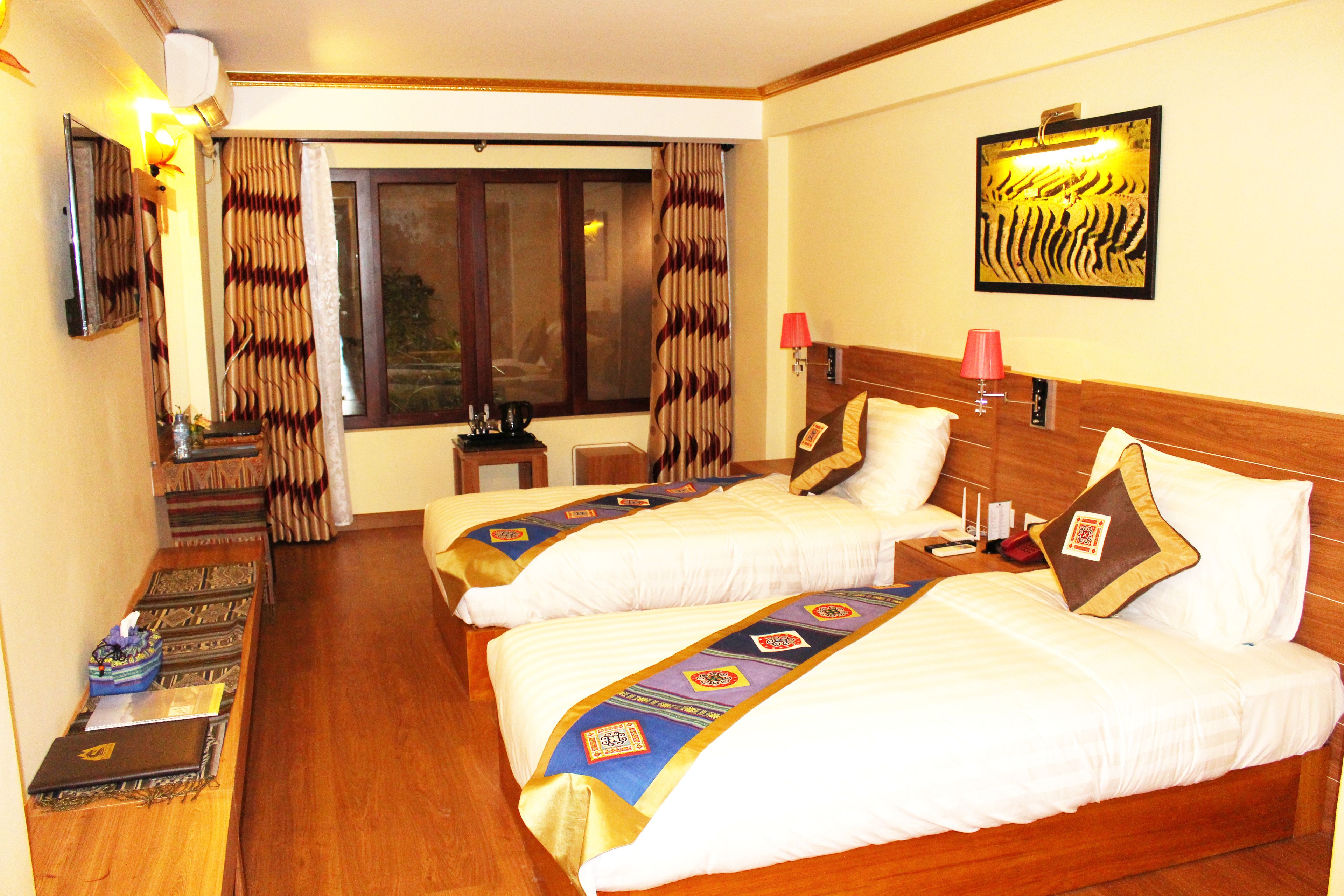 A hotel room with two beds, a desk, a chair, a window with curtains, a wall-mounted lamp, and a framed artwork.