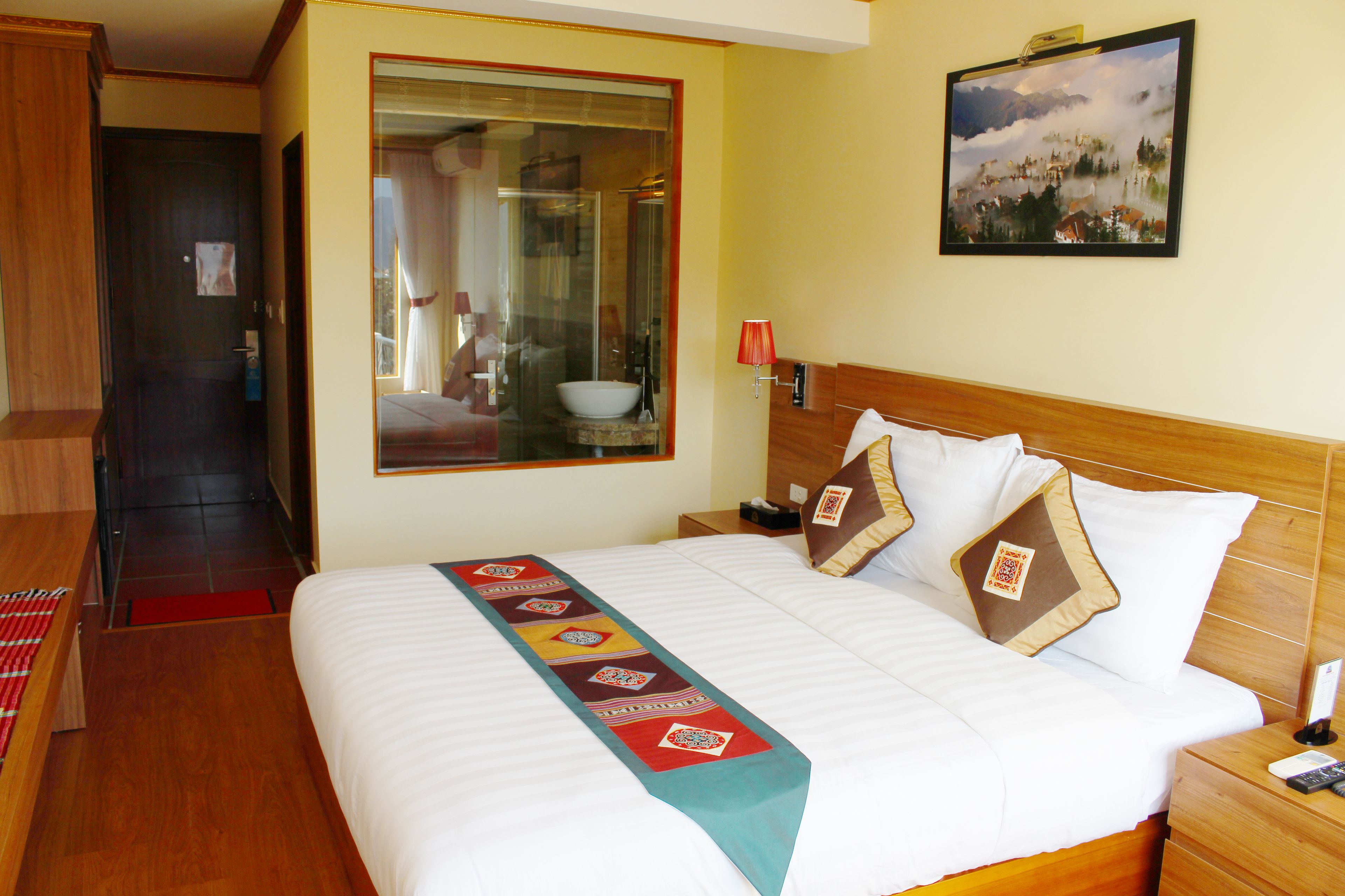 Una camera d'hotel con un letto grande, una testiera in legno, un comodino con una lampada e una finestra vista paesaggio montano.