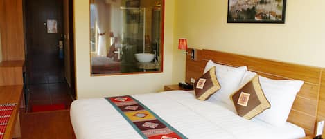 A hotel room with a large bed, a wooden headboard, a bedside table with a lamp, and a window with a view of a mountain landscape.