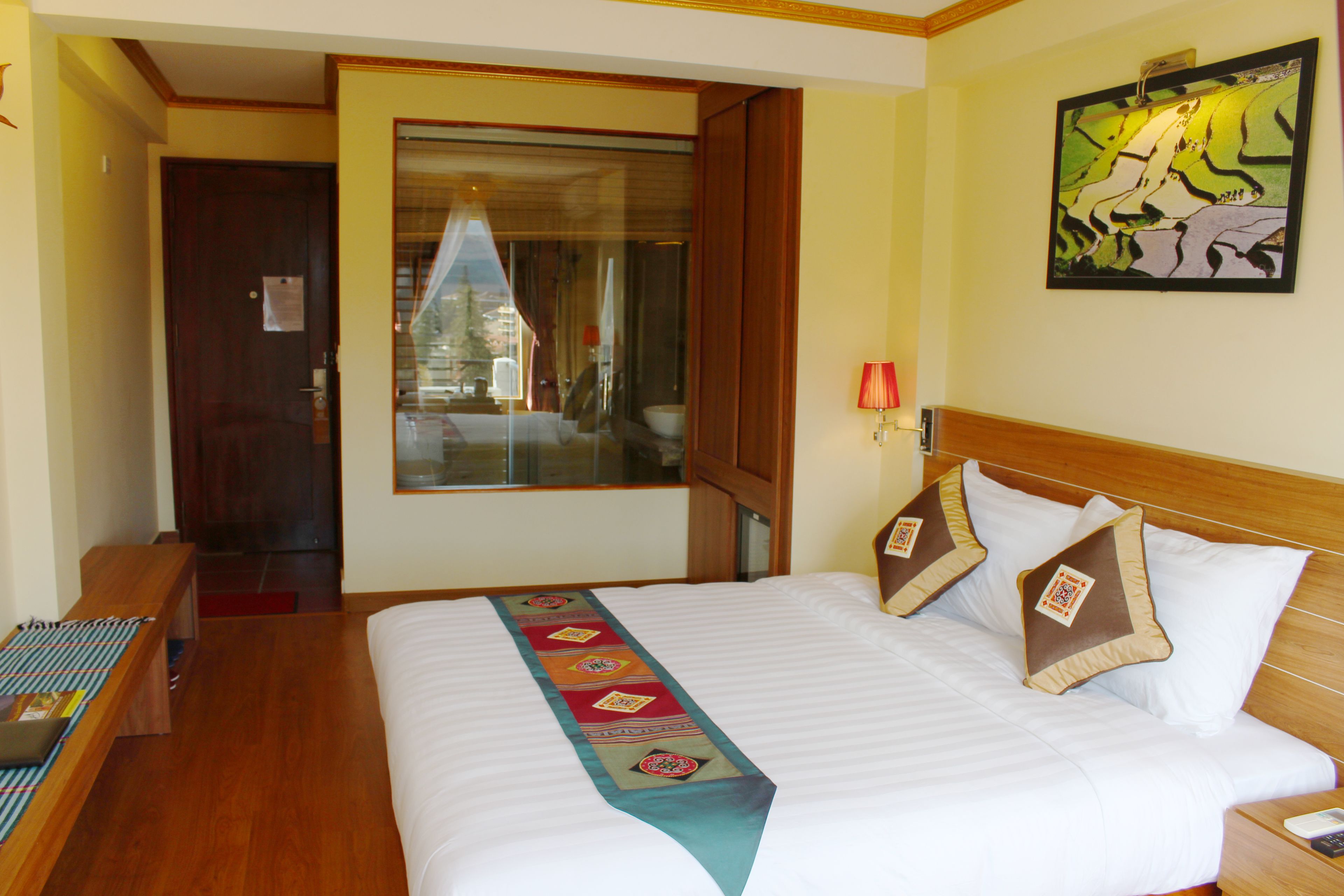 Quarto de hotel com uma cama grande, cabeceira de madeira, janela com vista e um quadro na parede.