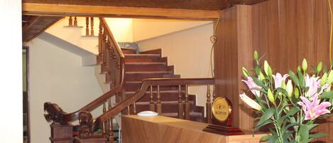 A wooden reception desk with a vase of flowers, a clock, and a staircase in the background.