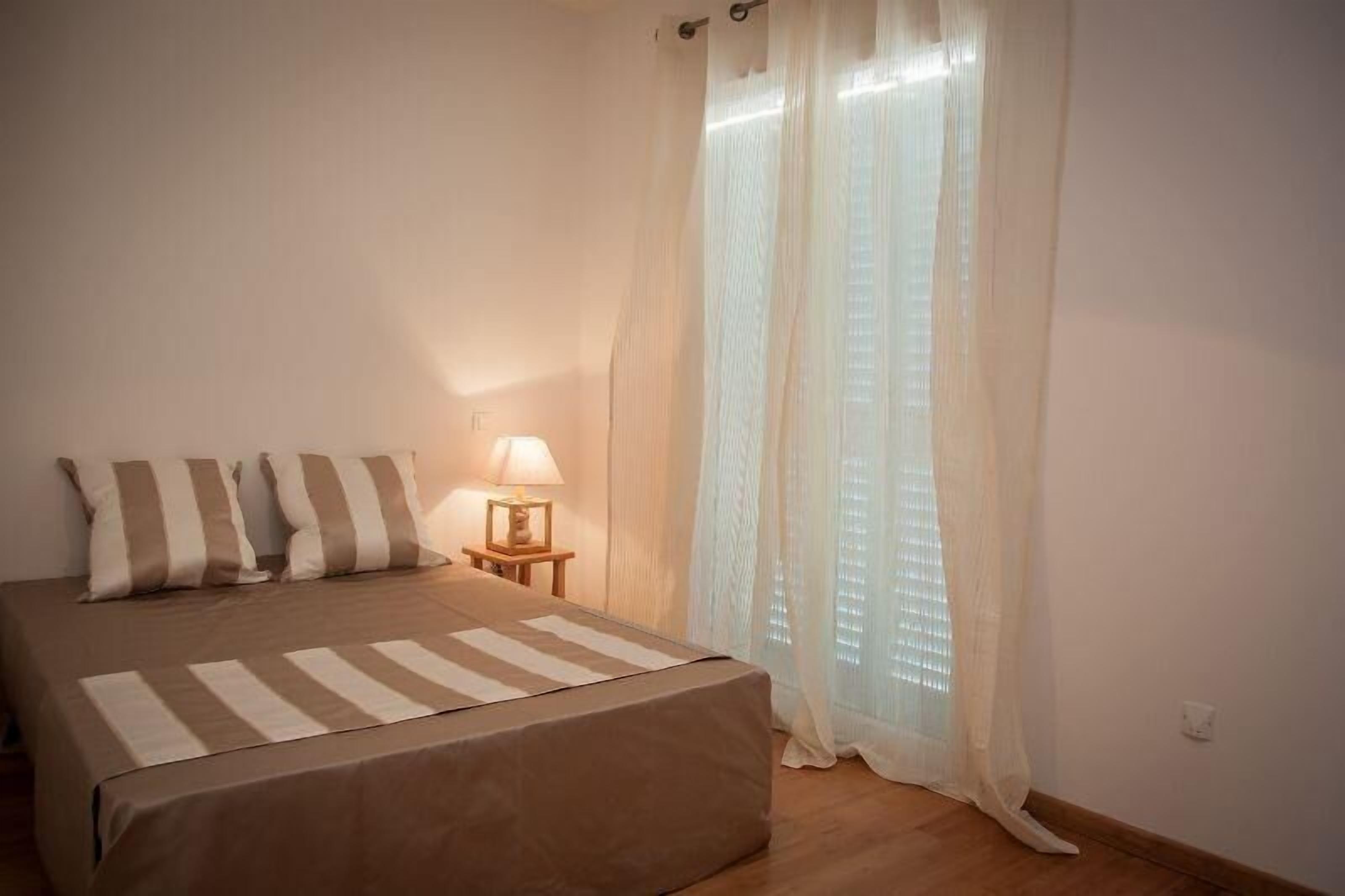 Un dormitorio con cama, almohadas, una mesita de noche con lámpara y cortinas translúcidas.