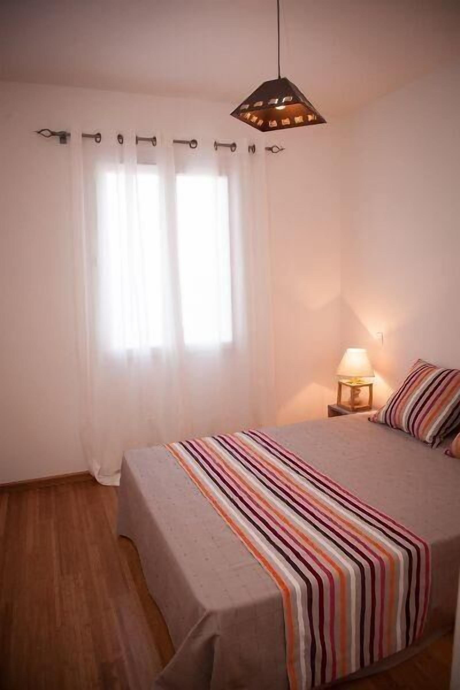 Quarto com cama, luminária de cabeceira, janela com cortinas e luminária pendente.