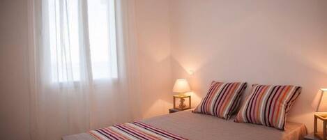 Ein Schlafzimmer mit einem Bett, zwei Nachttischlampen, einem Fenster mit durchsichtigen Vorhängen und einer gestreiften Decke.