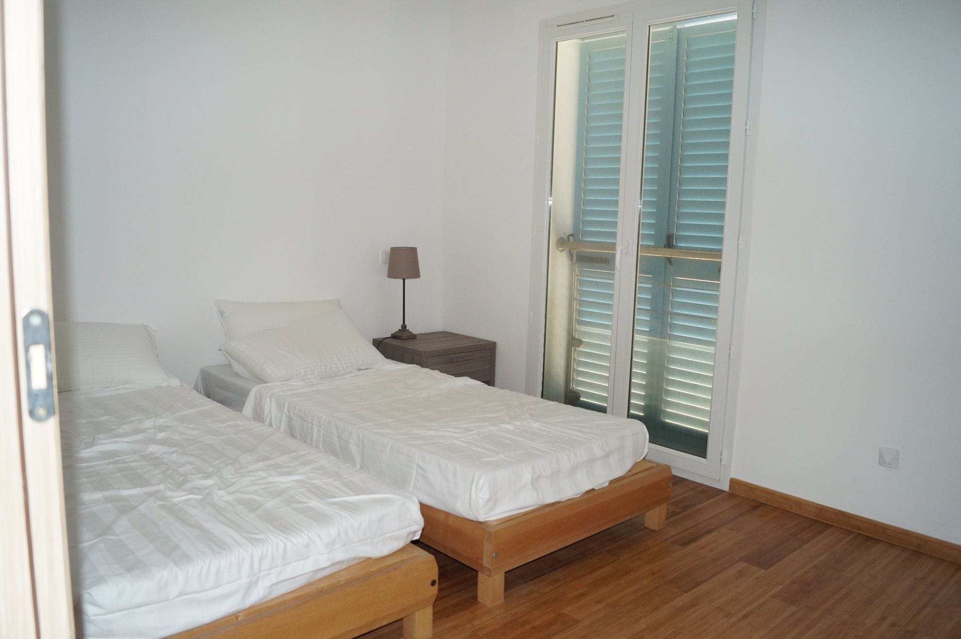 A bedroom with two single beds, a nightstand with a lamp, and a sliding glass door with blinds.