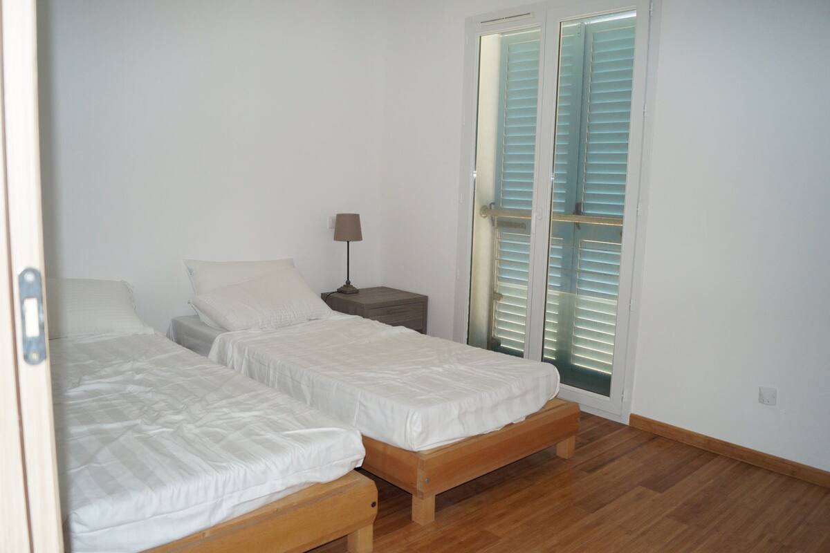 Quarto com duas camas de solteiro, mesa de cabeceira com abajur e porta de vidro deslizante com persianas.
