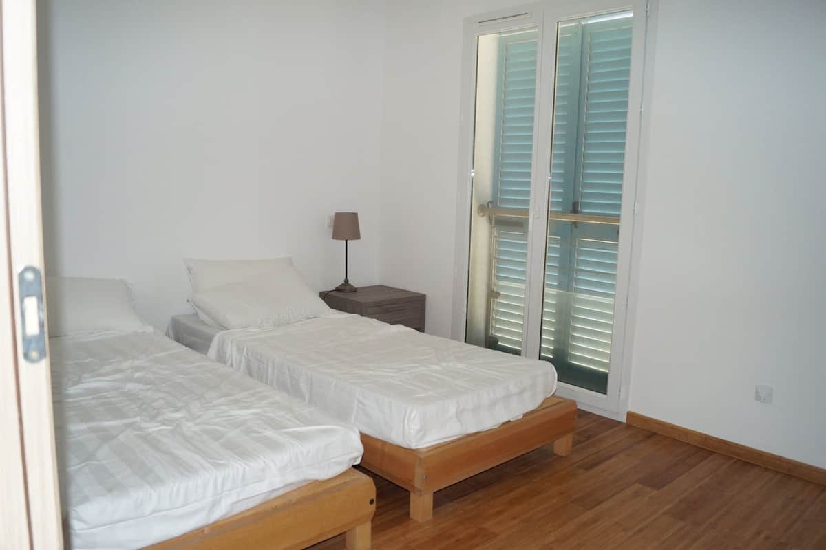 A bedroom with two single beds, a nightstand with a lamp, and a sliding glass door with blinds.