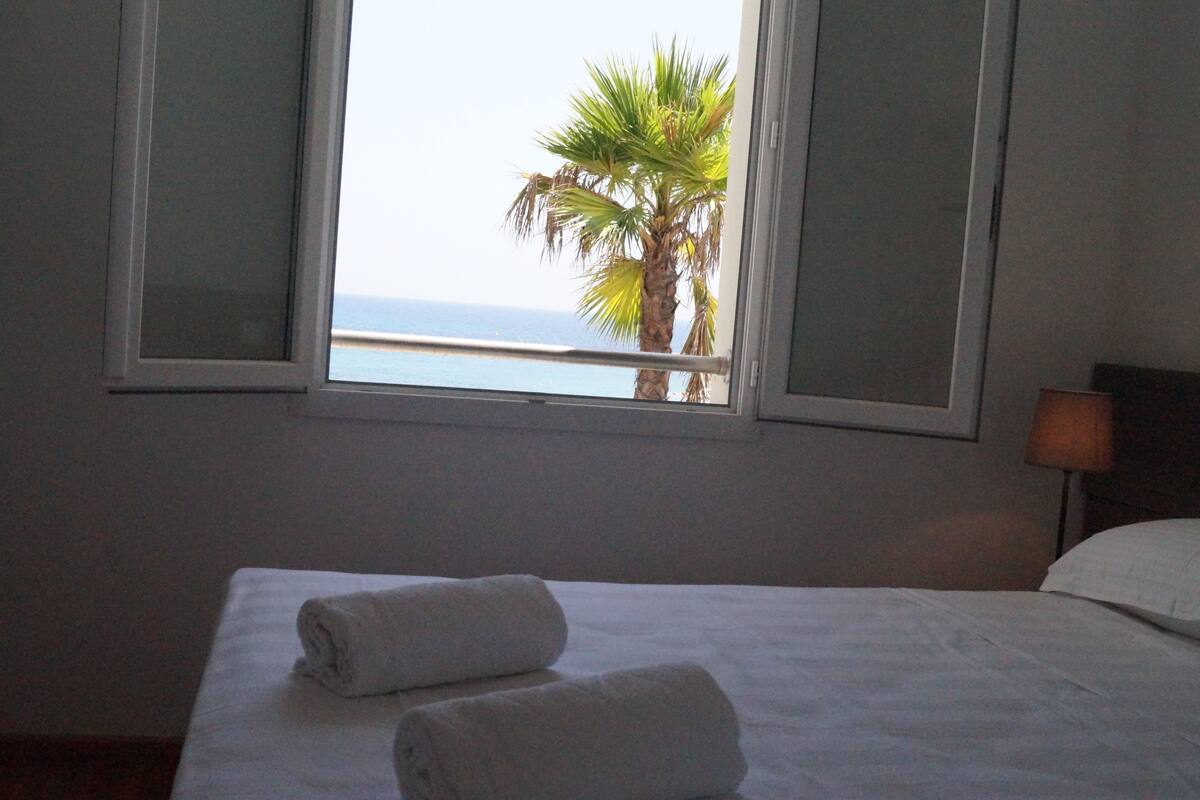 Quarto de hotel com cama, duas toalhas enroladas, janela com vista para o mar e uma palmeira, e mesa de cabeceira com abajur.