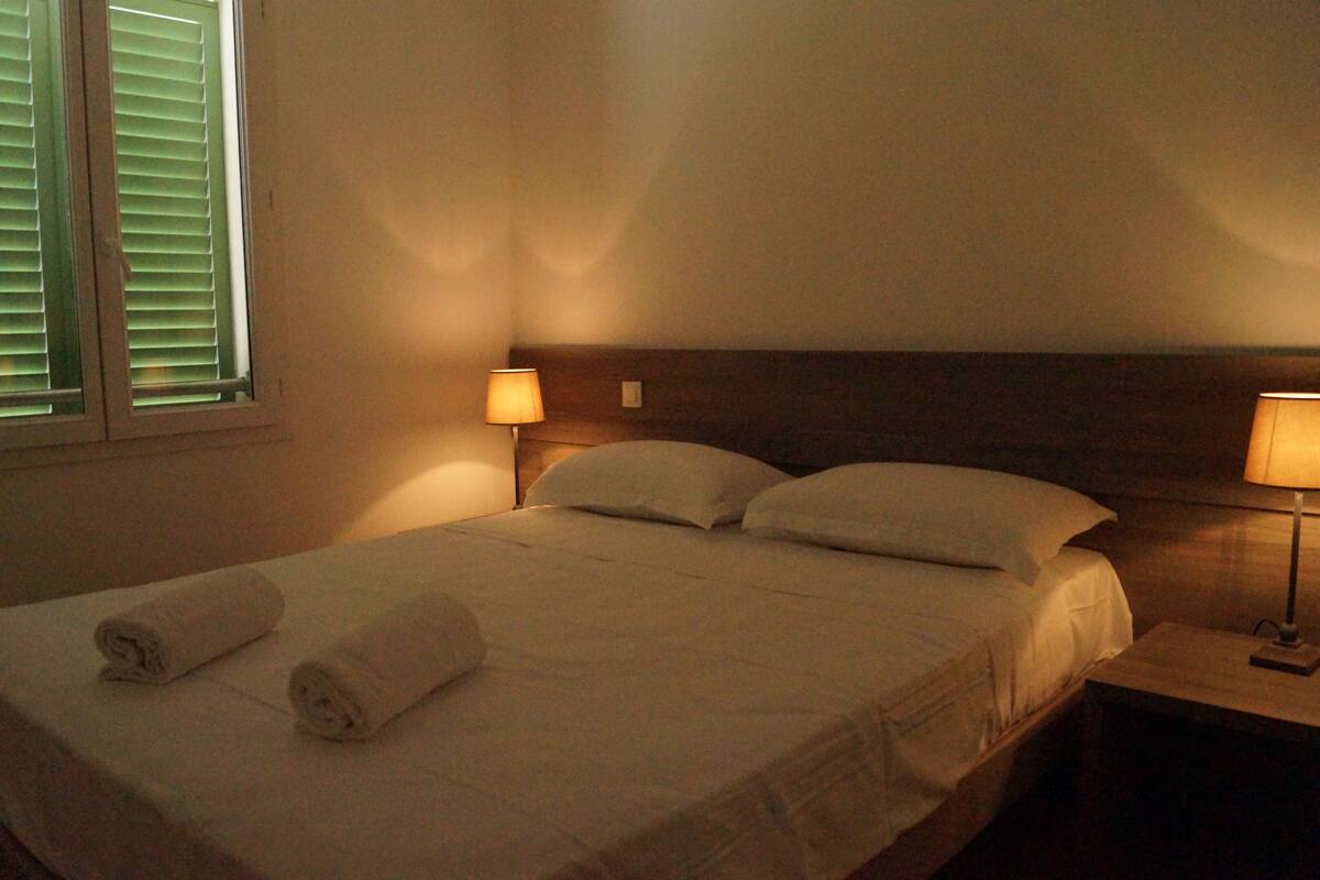 Cama bem arrumada com duas toalhas enroladas, cabeceira de madeira e duas luminárias laterais.
