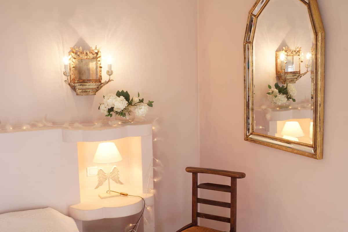 Een slaapkamer met een wit bed, een nachtkastje met lamp, een spiegel en een kleine houten stoel.