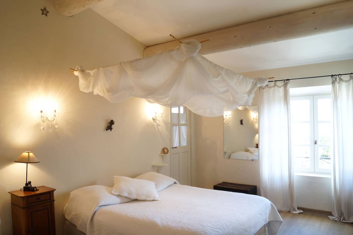 Een slaapkamer met een hemelbed, nachtlampjes, een nachtkastje en een raam met gordijnen.