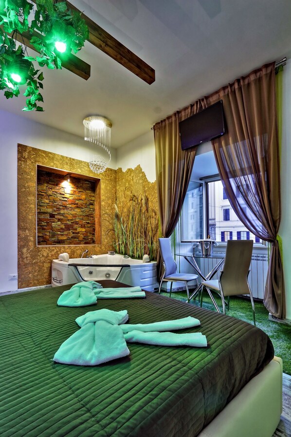 Une chambre d’hôtel avec un lit aux couleurs vertes, un mur en pierre, un plafond en bois, une télévision, une fenêtre avec des rideaux et un balcon avec une table et des chaises.