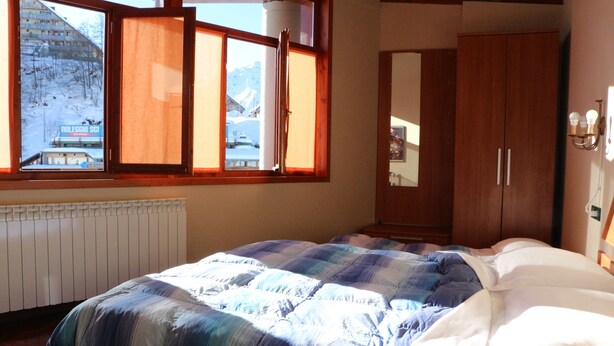 A bedroom with a bed, a window with a view of a snowy landscape, and a wooden wardrobe. - Hotel Laghetto (Frabosa Sottana)