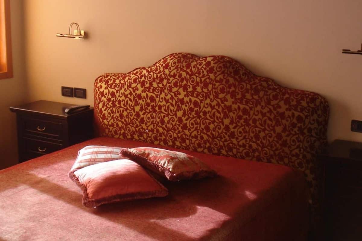 A hotel room with a patterned headboard, a bed with a red bedspread, and a nightstand with a lamp.