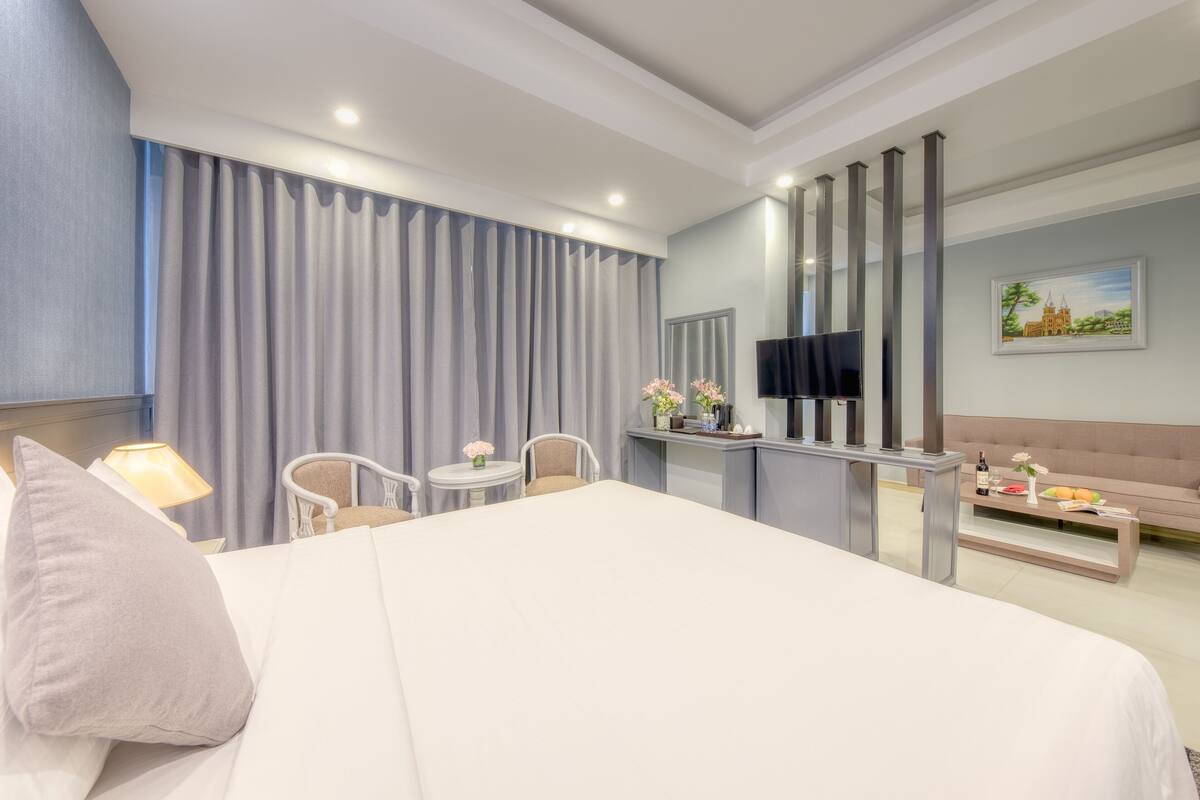 A hotel room with a large bed, a desk with a flat-screen TV, a sofa, and a dining table with a vase of flowers.