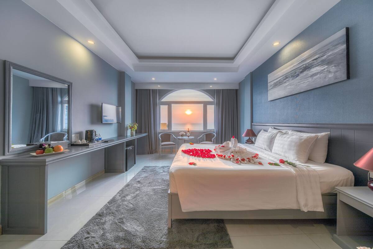 A modern hotel room with a large bed, a desk, a mirror, and a view of the sunset through the window.