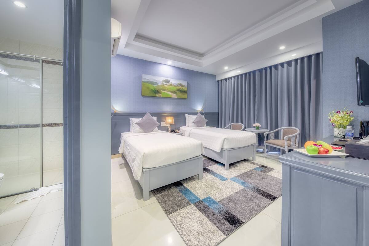 A modern hotel room with two beds, a shower area, a small table with a chair, and a TV mounted on the wall.