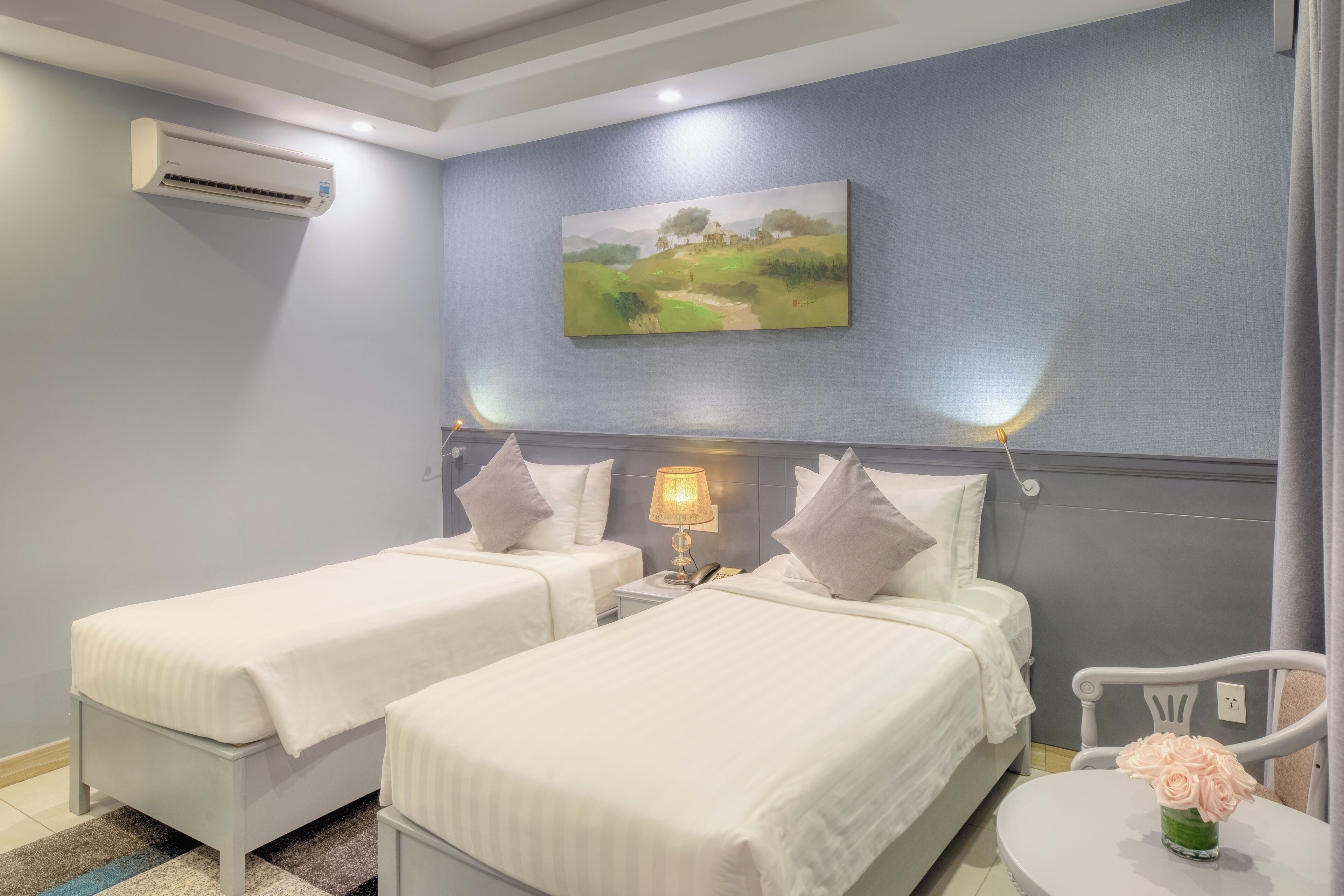 Una camera d'albergo con due letti singoli, un comodino con una lampada, un tavolino con un vaso di fiori e un quadro appeso al muro.