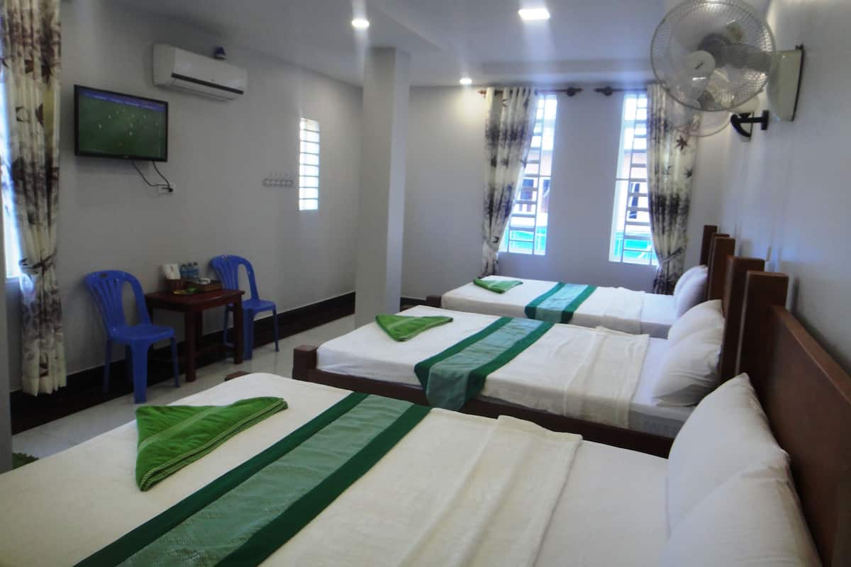 A hotel room with three beds, a desk with a chair, a TV mounted on the wall, and a window with curtains.
