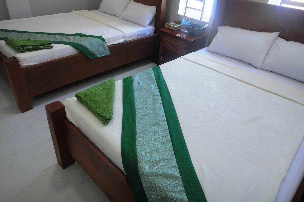 Two wooden beds with white linens and green accents, a wooden nightstand with a lamp, and a window with natural light.