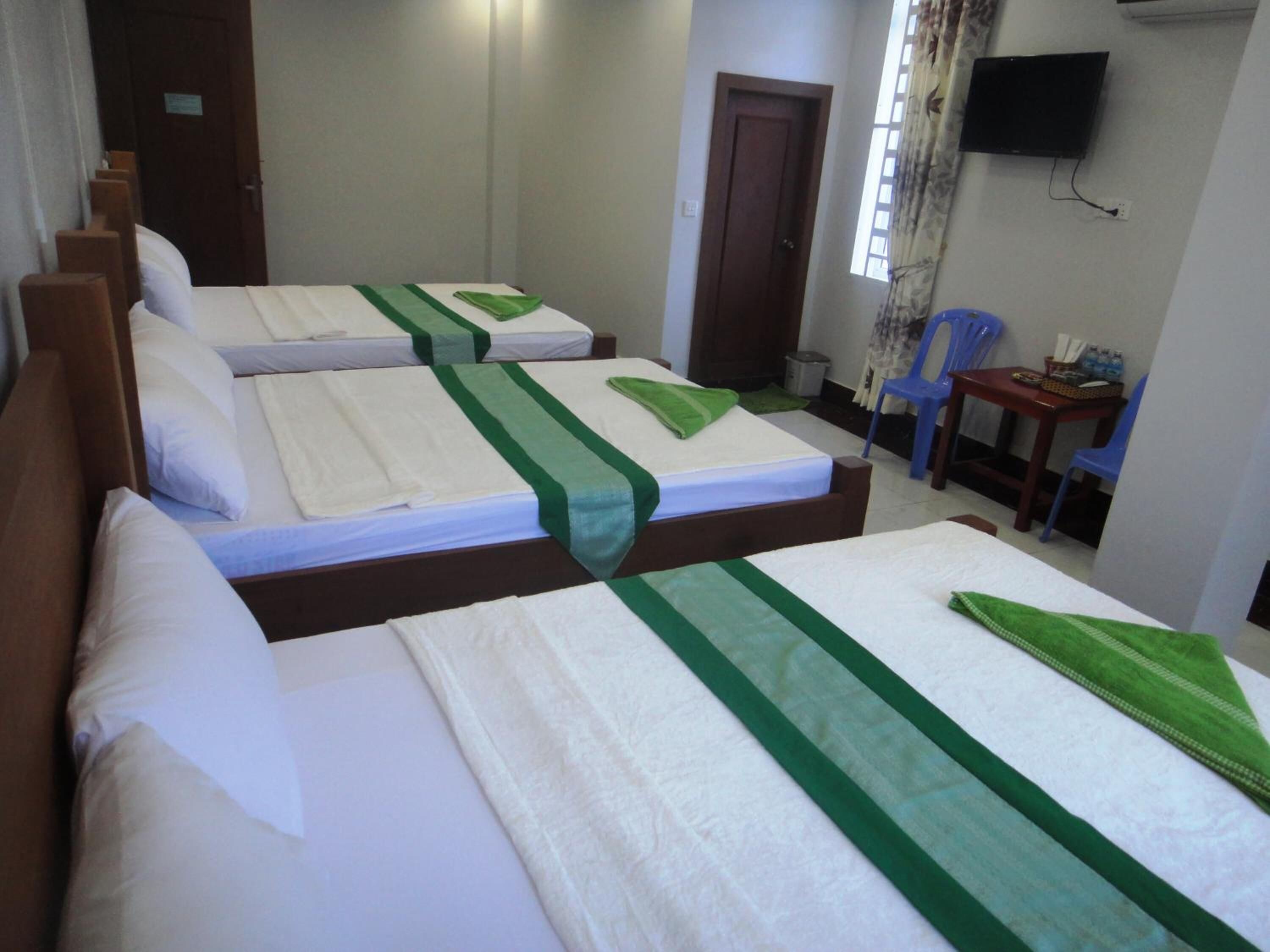 A hotel room with three single beds, each with a green bedspread and white linens. There is a wooden headboard, a small table with a chair, and a television mounted on the wall.