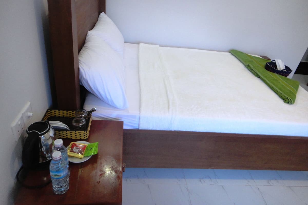 A neatly made bed with white linens, a wooden headboard, and a green towel on the side. A small table beside the bed holds a coffee maker, a bottle of water, and a packet of snacks.