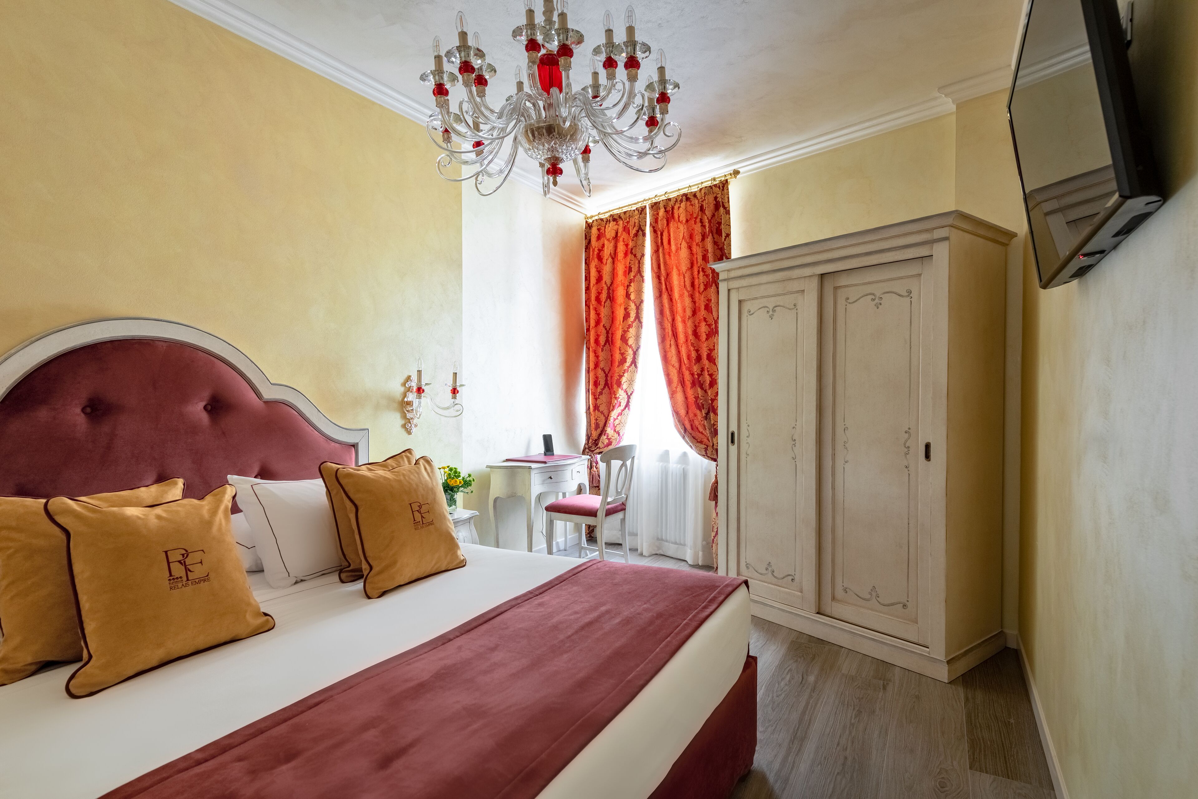 A hotel room with a large bed, a chandelier, a wardrobe, and a desk with a chair.