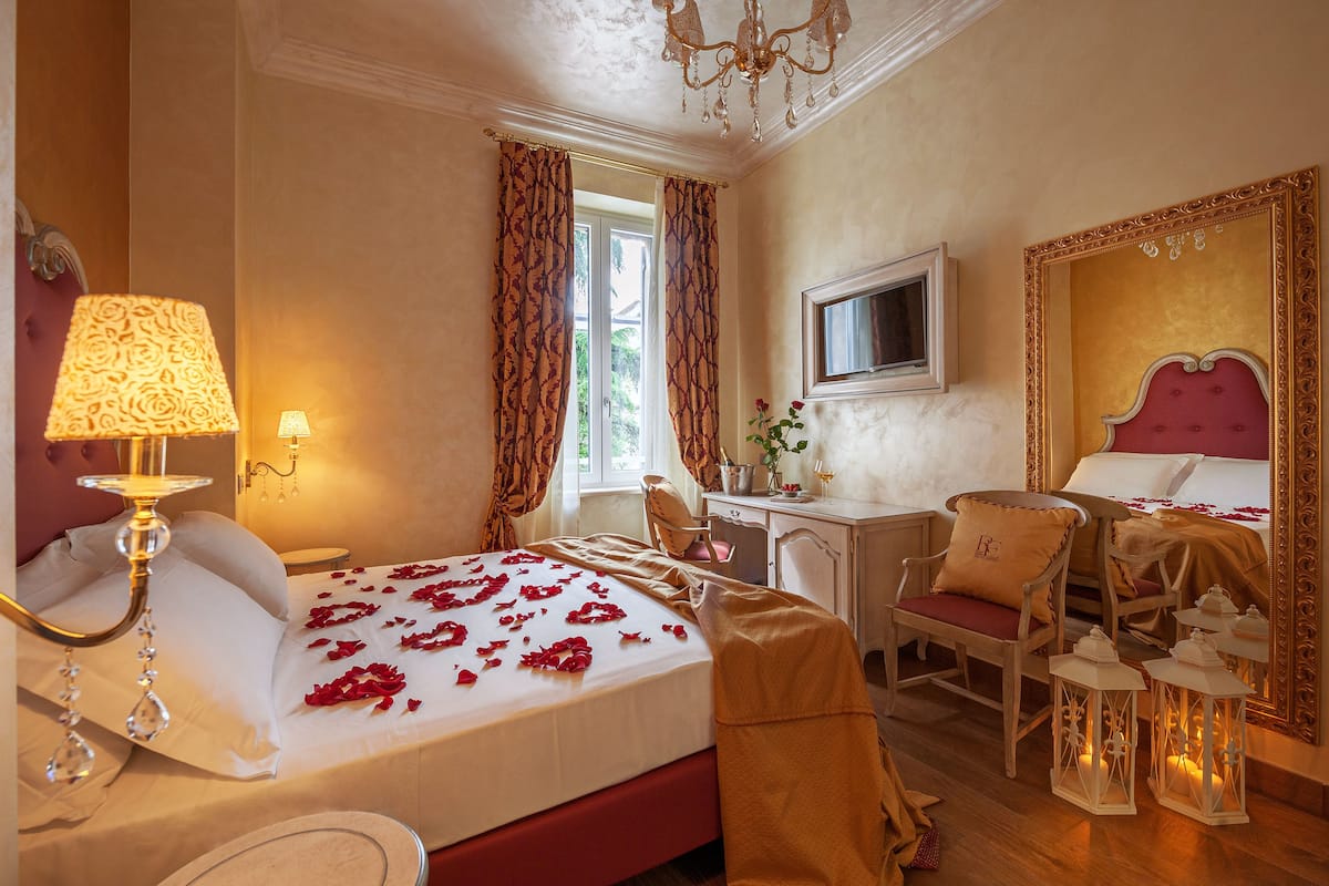 A hotel room with a bed, a desk, a chair, a chandelier, and a large mirror.