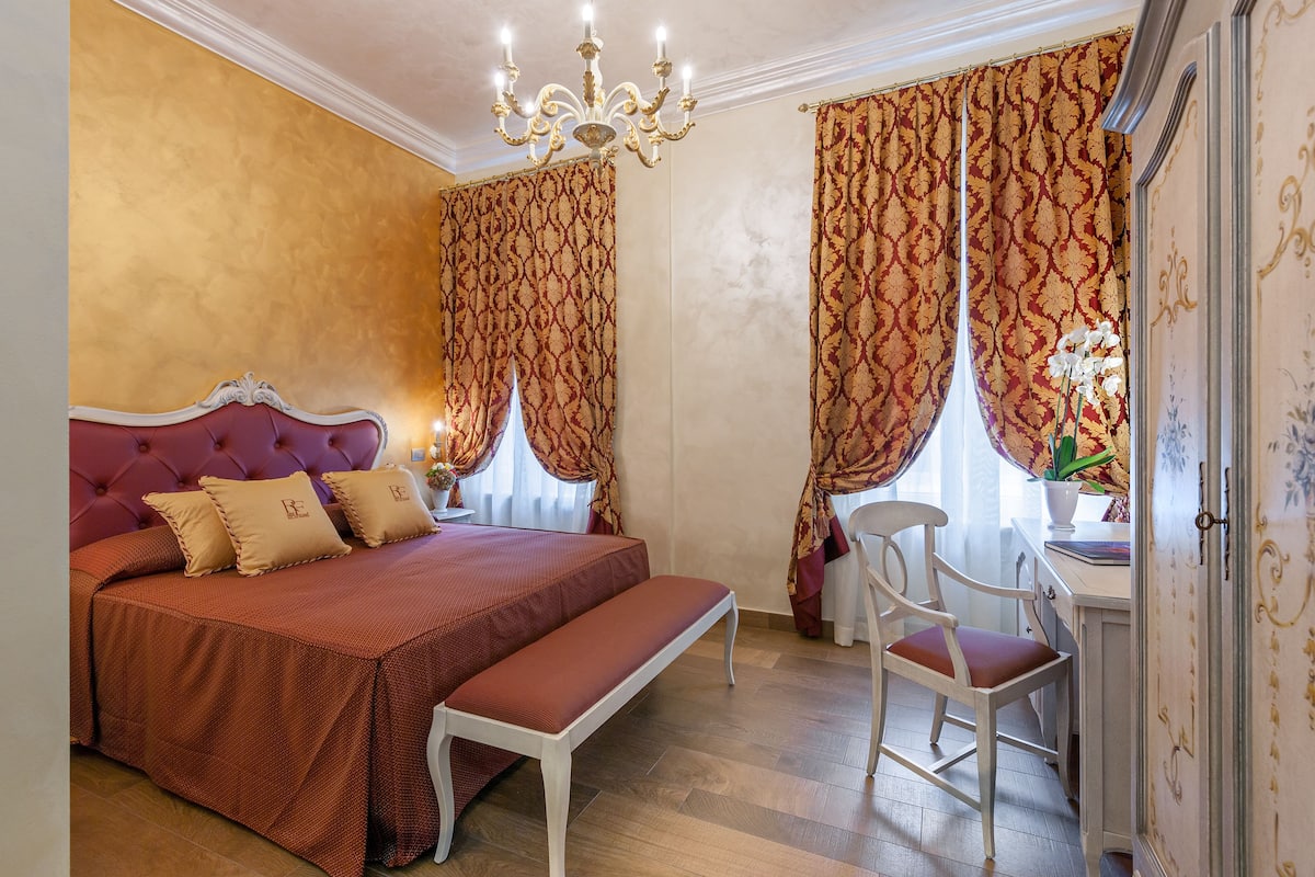 A bedroom with a large bed, a bench, a chair, and a desk with a vase of flowers.
