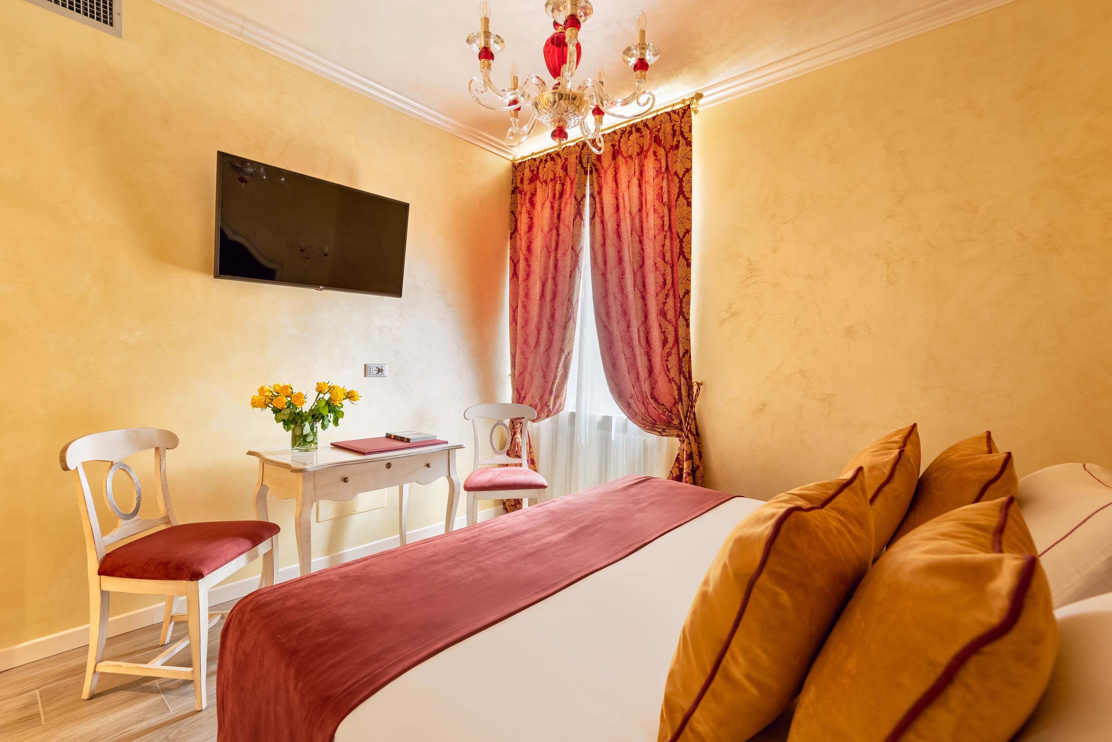 A hotel room with a bed, a desk, a chair, a TV mounted on the wall, and a window with curtains.