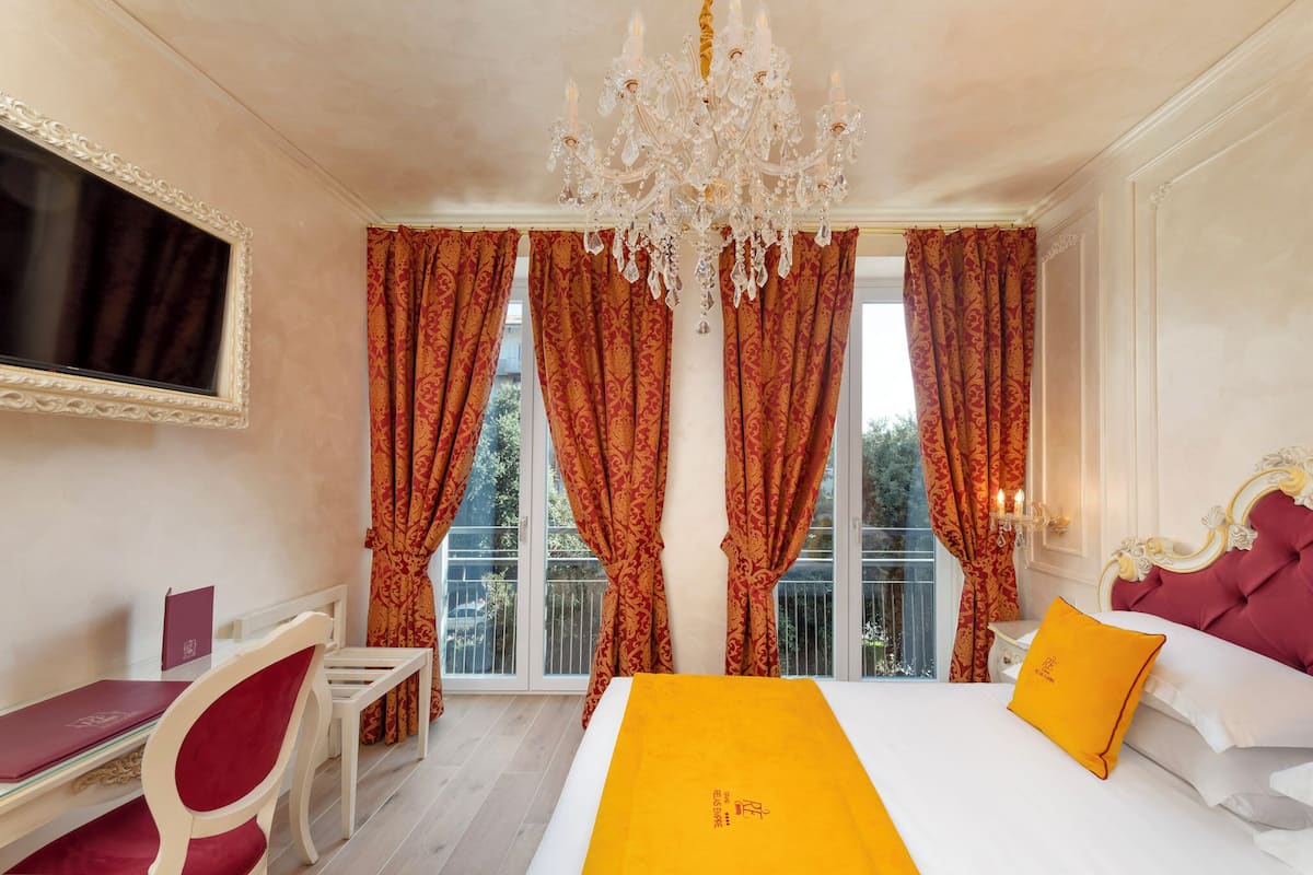 A hotel room with a large bed, a desk with a chair, a chandelier, and red curtains.